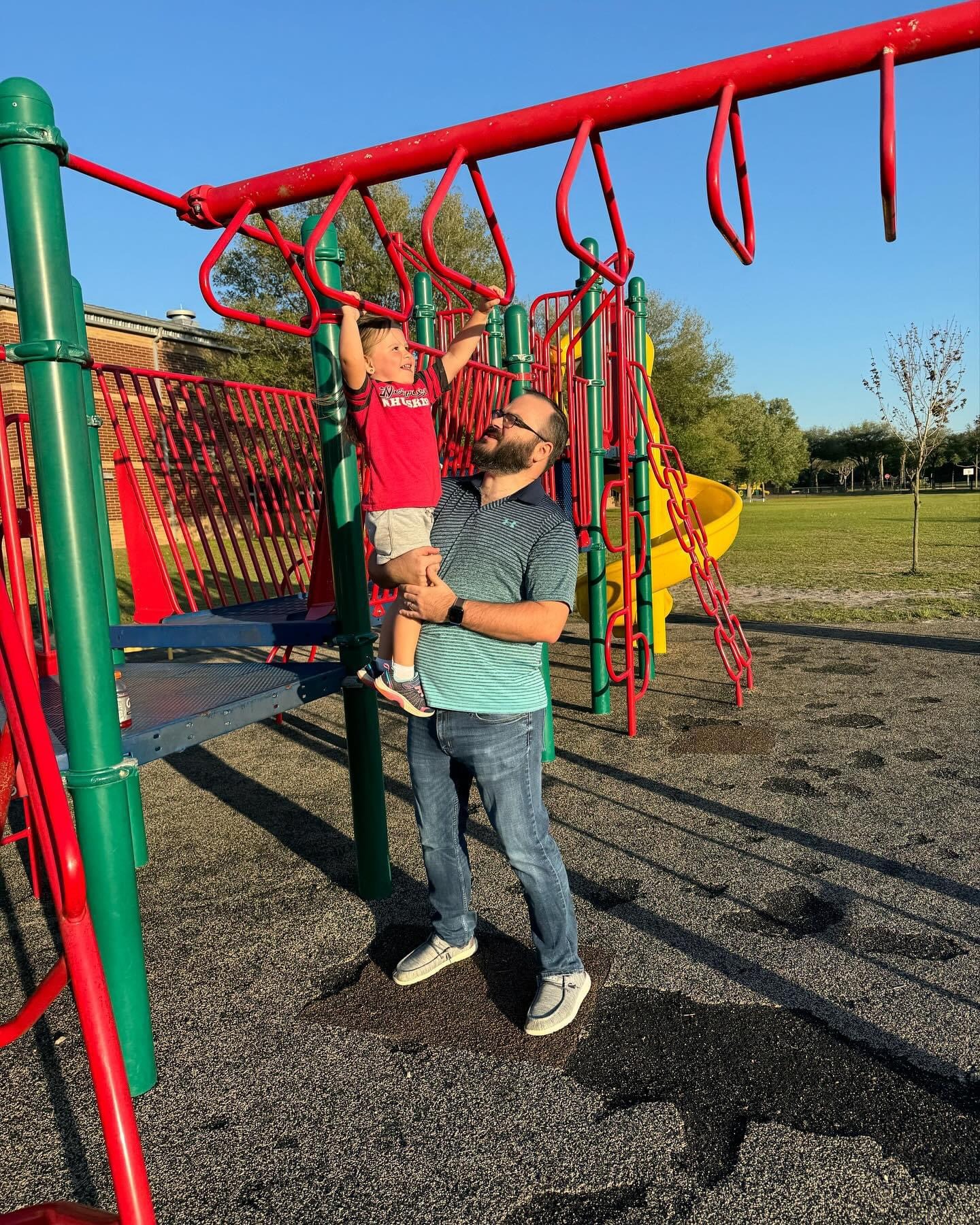 Michael supports his daughter as she reaches for the monkey bars at a playground.