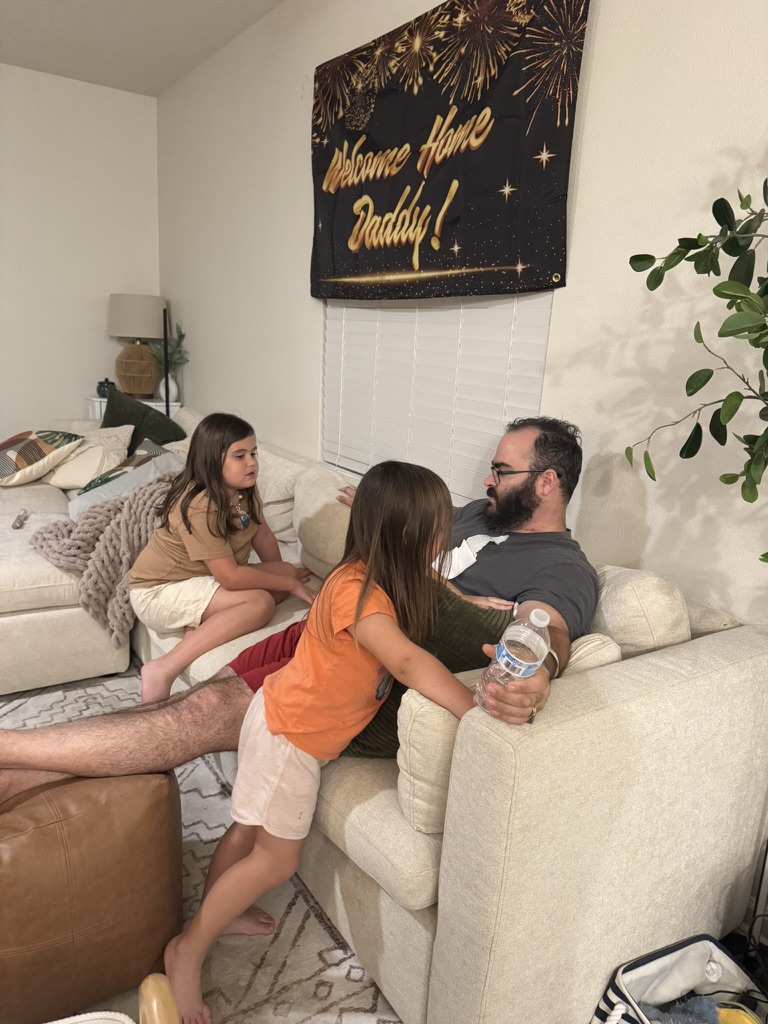 Michael rests on the couch with his daughters beneath a Welcome Home Daddy banner.