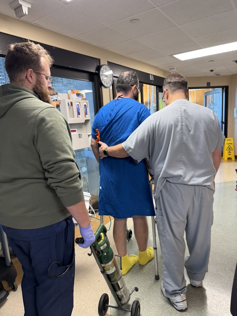 Michael uses a walker in a hospital hallway with staff alongside him and a portable oxygen cylinder.